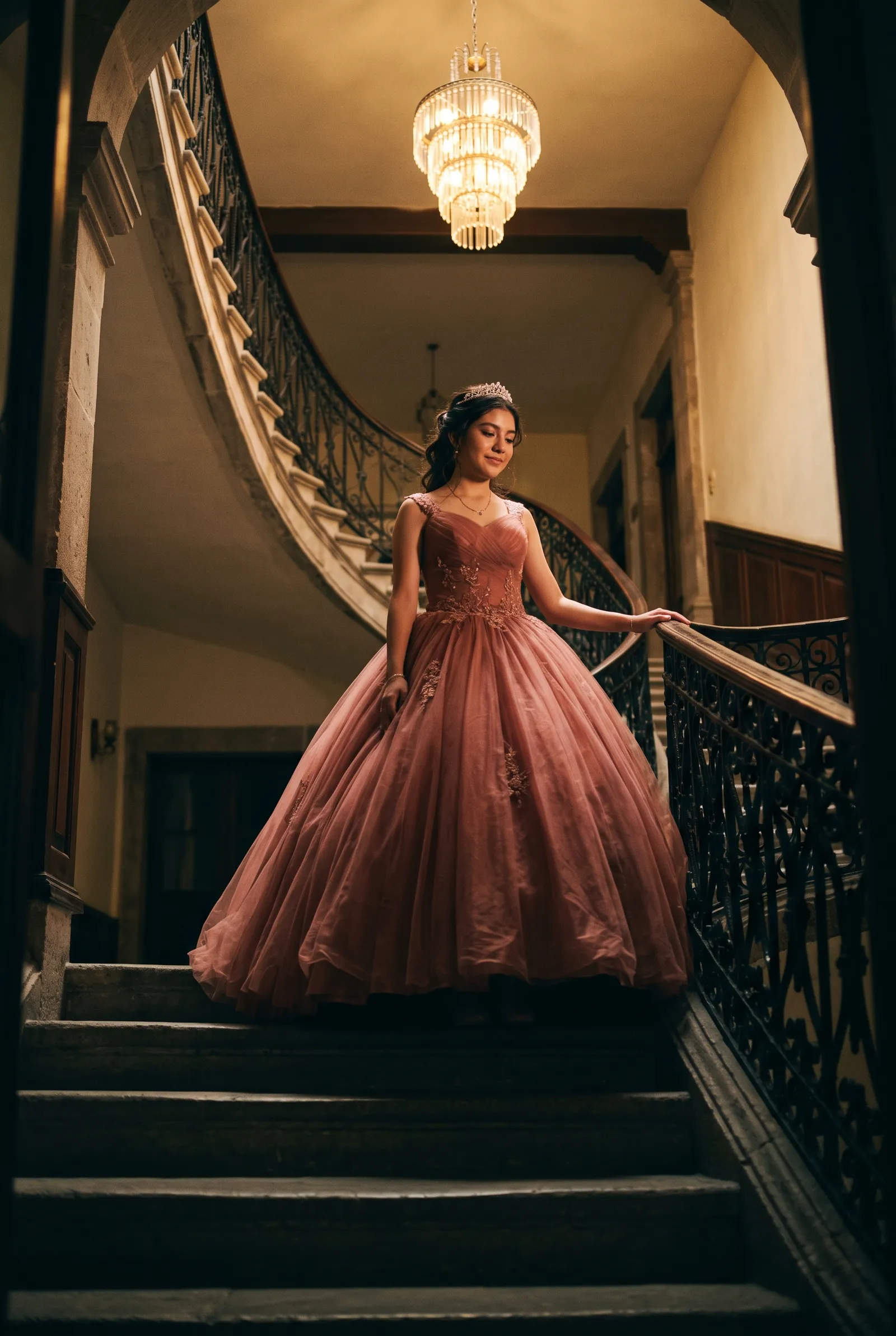 Quinceañera con vestido de tul en lo alto de una escalera colonial bajo un candelabro